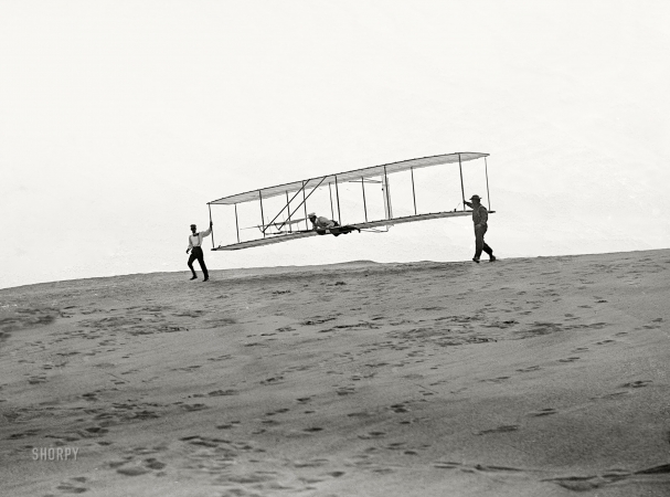 Photo showing: Testing Their Wings -- Oct. 10, 1902. Kitty Hawk, North Carolina. Start of a glide; Wilbur in motion
at left holding one end of glider, Orville lying prone in machine, and Dan Tate at right.