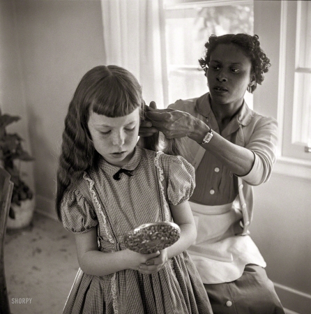 Photo showing: Helping Hands -- February 1956. Domestic worker fixing girl's hair.