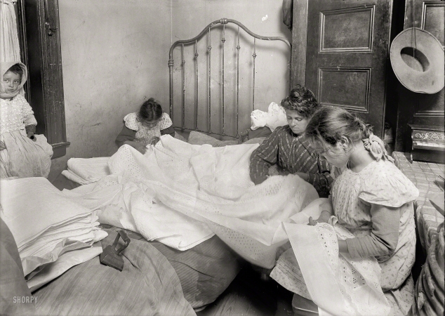 Photo showing: Cutting Bee -- November 1912. New York. Family cutting embroidery in tiny crowded bed-room.