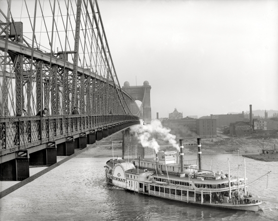 Photo showing: Steam Under the Bridge -- Circa 1906. The sidewheeler Cincinnati passing under the Roebling Suspension Bridge on the Ohio River. 