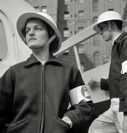 Photo showing: In a Jiffy -- November 1942. Hospital preparations for air raids involve the training of street accident units
for increased efficiency. Nurses wear coat and skirt that can be put on over uniform in a jiffy.