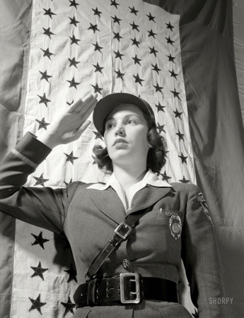 Photo showing: The Correct Salute -- March 1943. Estelle Hendel, a guard at the Bendix Aviation Plant,
stands before the company service flag and gives the correct salute.