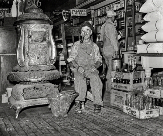 Photo showing: The Hot Seat -- October 1939. Farmer sits near the stove. General store in Lamoille, Iowa.