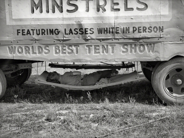 Photo showing: Rest Stop - -- May 1938. Carnival employee sleeping in hammock underneath wagon of Lasses White show. Sikeston, Missouri.