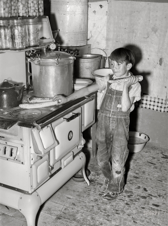 Photo showing: My Ears Are Burning -- July 1940. Spanish-American boy eating sweet corn roasted on top of hot stove. Chamisal, New Mexico.
