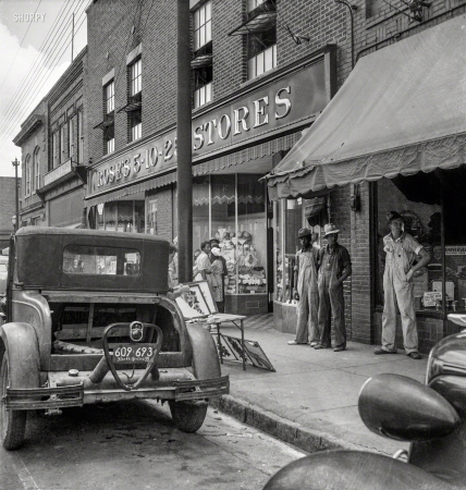 Photo showing: Five and Dime -- July 1939. Appliqued embroideries for sale on street in front
of ten cent store. Saturday afternoon. Siler City, North Carolina. 