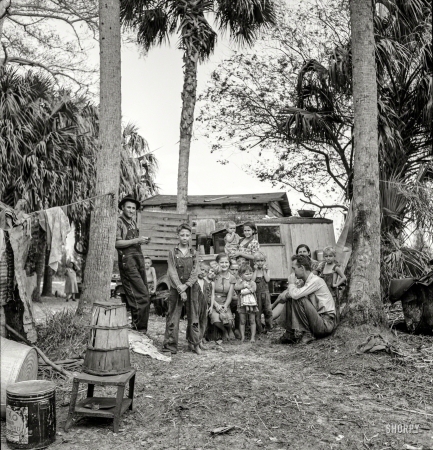Photo showing: Muck Rockers -- February 1939. Migrant laborers' camp near Canal Point, Florida. In foreground
is a bean hamper which they use to sit on and they call a 'muck rocker'.