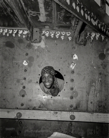 Photo showing: Baltimore, U.S.A. -- May 1943. Baltimore, Maryland. Building the SS Frederick Douglass.
Smiling from porthole of the dockhouse is rivet heater Willie Smith.