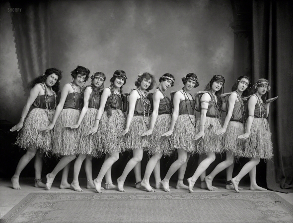 Photo showing: Lasses in Grasses -- New Zealand circa 1920s. Row of young women (music hall dancers?) in costume,
barefoot in dance pose, with headbands, beads, Hawaiian hula style grass skirts and anklets. 