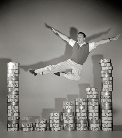 Photo showing: Over My Oeuvre -- 1952. Entertainer Gene Kelly executing leap over film cans labeled with titles of his movies.