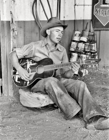 Photo showing: The Texaco Troubadour -- June 1940. Pie Town, New Mexico. Farm boy playing guitar in front of the filling station and garage.