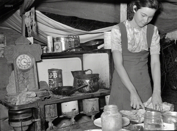 Photo showing: Homeless Cooking -- February 1939. Kitchen table and stove of white migrant tent camp near
Harlingen, Texas. Married daughter of migrant worker cutting salt meat for dinner.