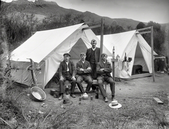 Photo showing: All the Comforts of Home -- Circa 1915. Four unidentified men in front of a tent, showing a washing basin, pots and
teapot with cups of tea, camping ground unidentified, possibly Christchurch district. 