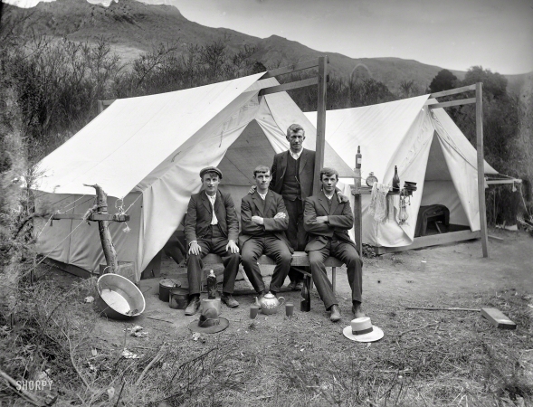 Photo showing: All the Comforts of Home -- Circa 1915. Four unidentified men in front of a tent, showing a washing basin, pots and
teapot with cups of tea, camping ground unidentified, possibly Christchurch district. 