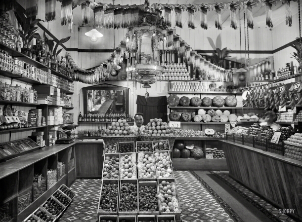 Photo showing: Apple Store -- Circa 1920s New Zealand. Greengrocery, probably Taranaki region. Chinese shopkeeper
with baskets of apples and boxes of peaches, cape gooseberries and other fruits. 